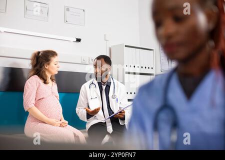 Kaukasische werdende Mutter sitzt auf dem Krankenhausbett, während ein männlicher Arzt im Labormantel sie durch den Plan zur pränatalen Versorgung spricht. Schwarze Krankenschwester prüft medizinische Akten, während der Arzt eine schwangere Patientin berät. Stockfoto