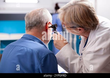 Pensionierter Mann, der im Krankenhauszimmer sitzt, unterzieht sich einem Gehörtest, während eine hochrangige hno-Fachärztin eine medizinische Untersuchung mit Otoskop durchführt. Älterer Arzt, der das Ohr männlicher Rentner in der Klinik untersucht. Stockfoto