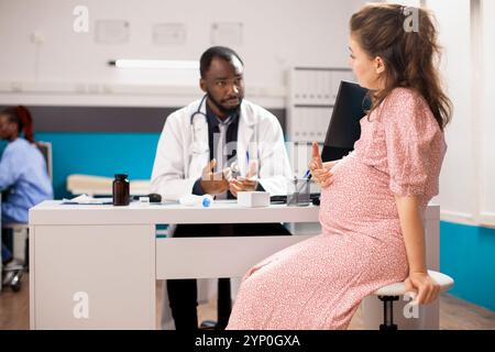 Die werdende Mutter spricht mit der afroamerikanischen Geburtshelferin über ihren Schwangerschaftsverlauf während der ärztlichen Untersuchung. Freundlicher Arzt, der schwangeren kaukasischen Frau zuhört, während er am Schreibtisch der Klinik sitzt. Stockfoto