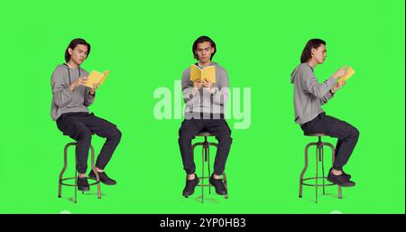 Trendige Person, die Romanbuch auf Stuhl vor Ganzkörper-Greenscreen-Vorlage liest und sich an Old School-Vorlesungshobbys interessiert. Junger asiatischer Erwachsener, der Literatur oder fiktionales Buch hält, um im Studio zu lesen. Stockfoto