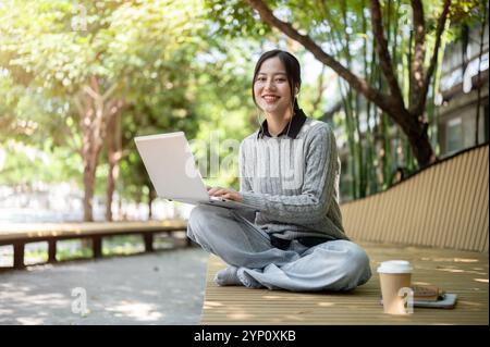 Eine charmante, glückliche asiatische Frau arbeitet im Freien, sitzt mit ihrem Laptop auf einer Parkbank, trägt Ohrhörer und lächelt in die Kamera. Stockfoto