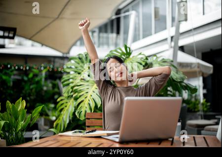 Eine entspannte, sorglose asiatische Frau streckt ihre Arme, während sie draußen vor ihrem Laptop sitzt und von einem Café in der Stadt aus ferngesteuert arbeitet Stockfoto