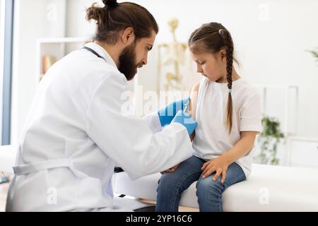 Männlicher Arzt geimpft junge kaukasische Mädchen auf medizinischer Couch mit Spritze. Der Arzt trägt einen weißen Mantel, Handschuhe und konzentriert sich auf den Arm des Mädchens. Szene zeigt Gesundheitswesen, medizinische Versorgung und Impfprozess. Stockfoto