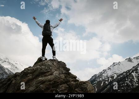 Rückansicht der Rückseite des jungen Wanderers, der mit Happy auf dem Gipfel des felsigen Berges steht und die Hände aufrichtet, Rucksacktouristen auf einem Berg mit ausgestreckten Armen Stockfoto