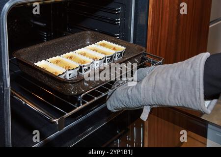 Eine Person, die einen Ofenhandschuh trägt, stellt ein Tablett mit ungekochten Desserts in einen vorgewärmten Ofen, um das Backen vorzubereiten. Stockfoto
