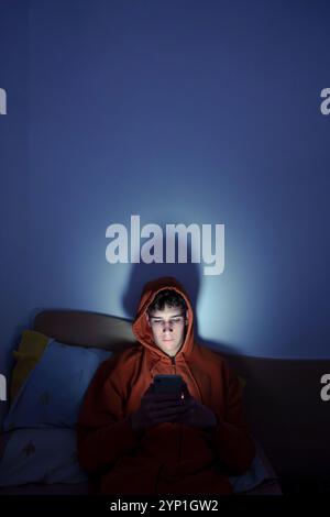 Teenager, der nachts auf dem Bett sitzt und von der Bildschirmbeleuchtung beleuchtet wird Stockfoto