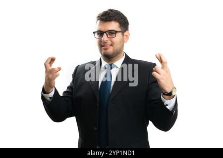 Hoffnungsvoller, erwachsener Unternehmer lächelnd in einem eleganten Anzug und einer Brille, die Glücksgeste mit gedrückten Daumen auf beiden Händen, isoliert auf Stockfoto