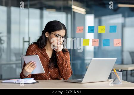 Hispanische Geschäftsfrau tief in Gedanken im modernen Büro mit Tablette. Schwerpunkt auf Notebook, umgeben von Haftnotizen, Telefon zur Hand, Darstellung der Arbeitsumgebung, Entschlossenheit, Multitasking. Stockfoto