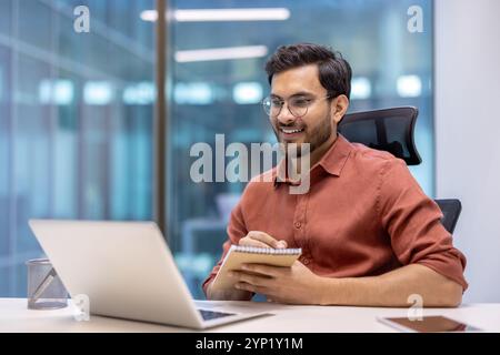 Ein selbstbewusster Mann, der an Online-Schulungen am Arbeitsplatz teilnimmt, lächelt den Laptop an, während er sich Notizen im Notebook macht. Repräsentiert Konzepte des Business Learning, der digitalen Interaktion, des technisch versierten Profis. Stockfoto