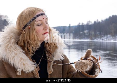 Eine junge Frau mit blonden Haaren, gekleidet in einem warmen Pelzmantel mit Accessoires im Boho-Stil, am Ufer an einem verschneiten Wintertag Stockfoto