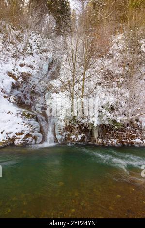 Kleiner Wasserfall im Winterwald. Malerische weiße Jahreszeit. Gefrorenes Wasser und blattlose Bäume auf dem schneebedeckten Hügel. Landschaft mit türkisfarbenem Bach Stockfoto