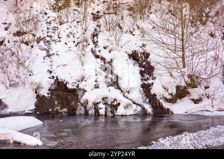 Kleiner Wasserfall im Winterwald. Eisbedeckter Bach. Gefrorenes Wasser und blattlose Bäume auf dem schneebedeckten Hügel. Abgelegenes Hinterland Stockfoto