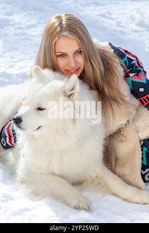 Eine schöne junge Blondine mit langen Haaren posiert im Park im Schnee mit einem weißen Huskyhund. Das Mädchen trägt einen warmen Strickpullover und ugg bo Stockfoto