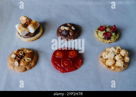 Hausgemachte runde Kekse auf Backpapier mit verschiedenen Belägen - Himbeere, Erdbeere, Pistazien, Karamell, Schokolade, Erdnüsse, Marshmallow, Popcorn. Stockfoto