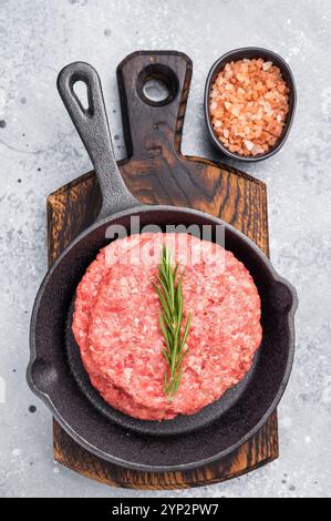 Fertig zum Kochen rohe Rindfleischpatties, Schnitzel mit Kräutern in der Pfanne. Grauer Hintergrund. Draufsicht. Stockfoto