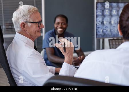 Seitenansicht eines erfahrenen männlichen Oberärzten in Gruppengesprächen mit Kollegen über die Behandlung während des Sitzens am Besprechungstisch in der Arztpraxis, c Stockfoto