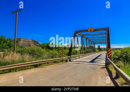 Eine Brücke überspannt eine Straße über ein grasbewachsenes Feld. Die Brücke ist alt und hat ein gelb-schwarzes Schild. Der Himmel ist blau und klar Stockfoto