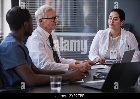 Klinikerin aus dem Mittleren Osten diskutiert Anamnese der Krankheit der Patienten mit Kollegen, die am Meeting-Tisch sitzen, während sie wichtige Punkte der Behandlung schreibt Stockfoto