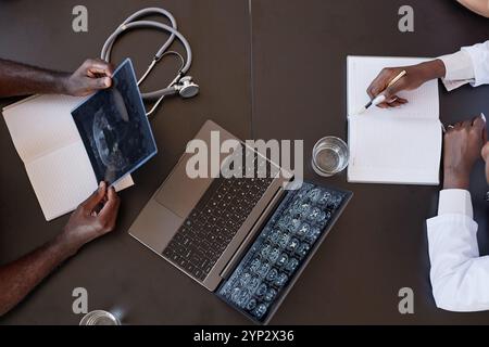 Draufsicht des Schwarzen Mannes, der einen Röntgenscan des Schädels hält, der am Tisch sitzt, mit einem Laptop, der CT-Bildgebung des menschlichen Gehirns auf dem Bildschirm anzeigt, während die Frau sich aufhält Stockfoto