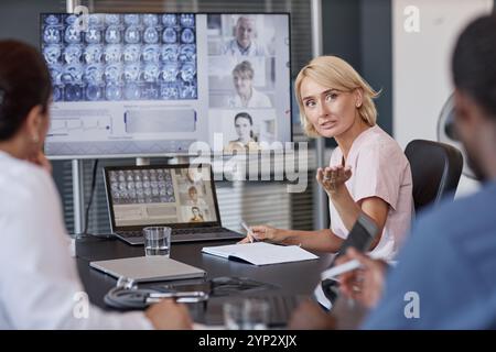 Radiologin präsentiert ihre Kollegin den Teilnehmern der Online-Konferenz, die mit einem Laptop am Meeting-Tisch sitzen, während sie gemeinsam über die Behandlung diskutieren Stockfoto