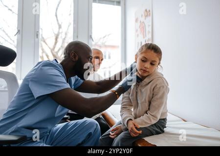 Männlicher medizinischer Experte, der das Ohr eines Mädchens überprüft, das im Untersuchungsraum im Krankenhaus auf dem Bett sitzt Stockfoto