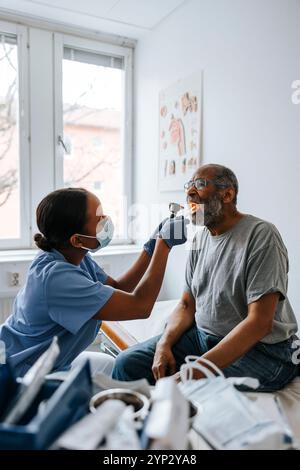 Kinderärztin untersucht den Hals eines älteren Mannes mit Zungendepressor und Taschenlampe im Untersuchungsraum Stockfoto