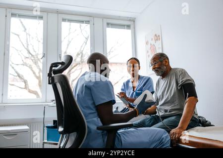 Männlicher Arzt, der den Blutdruck eines älteren Mannes überprüft, der neben der Krankenschwester auf dem Bett im Untersuchungsraum der Klinik sitzt Stockfoto
