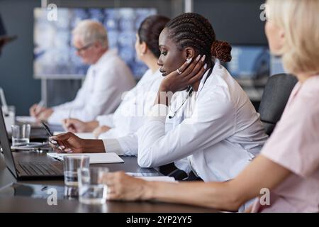 Seitenansicht einer afroamerikanischen Ärztin, die der Präsentation der Kollegen zuhört und Aufzeichnungen über Arbeitssitzungen hält, während sie mit Ärztin am Tisch sitzt Stockfoto