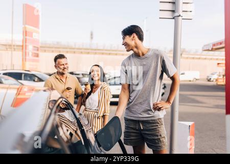 Glücklicher junger Mann, der das Auto mit dem Elektrostecker auflädt, während er mit den Eltern am Bahnhof redet Stockfoto