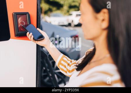 Über-Schulter-Sicht einer reiferen Frau, die mobiles Bezahlen an einer Elektrofahrzeugladestation tätigt Stockfoto