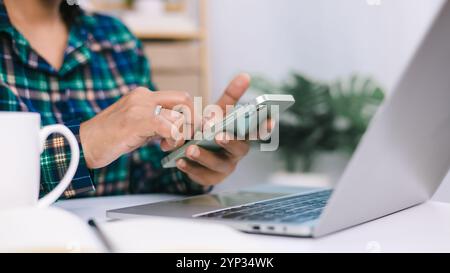 Eine Frau schaut auf ihr Handy, während sie an einem Schreibtisch sitzt. Sie konzentriert sich auf ihr Telefon und liest E-Mails oder Nachrichten. Produktivitäts- und Arbeitsauftragskonzept Stockfoto