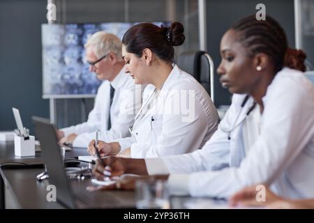 Seitenansicht einer Klinikerin aus dem Nahen Osten, die Kollegen-Präsentation hört und Aufzeichnungen schreibt, während sie am Meeting-Tisch in der medizinischen Abteilung sitzt Stockfoto