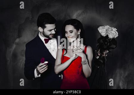 Porträt eines stilvollen Gentlemans mit Stoppeln, der einer aufgeregten, fröhlichen Dame einen goldenen Ring mit Diamanten gibt, mit Schmuck, der weiße Rosen in den Händen hält, Paar Stockfoto