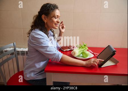 Eine Frau in einem blauen Hemd benutzt ein Tablett in ihrer Küche, um Kochrezepte zu erforschen. Frisches Gemüse ist in der Nähe, was einen Fokus auf gesunde Ernährung und Stockfoto