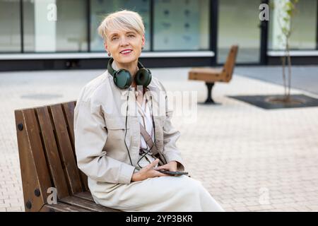 Schöne lächelnde reife Frau mit kurzen Haaren, die Kopfhörer trägt und das Telefon benutzt, während sie draußen auf der Bank in der Stadt sitzt, aktiver Lebensstil für ältere Stockfoto