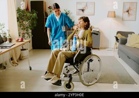 Betreuungsperson hilft älteren Frauen im Rollstuhl in komfortablem Wohnzimmer mit moderner Einrichtung und Pflanzen im Innenbereich Stockfoto