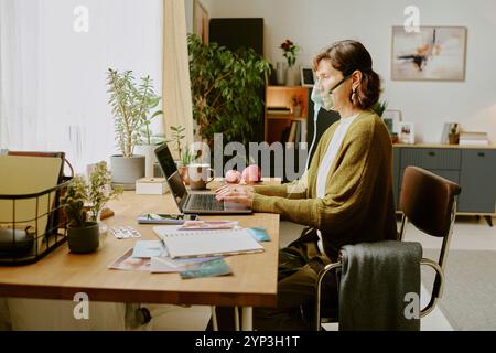 Ältere Frau mit Gesichtsmaske, die am Schreibtisch sitzt und im Heimbüro an einem Laptop arbeitet, umgeben von Pflanzen und Büchern, und während der Pandemie Sicherheitsmaßnahmen aufrechterhält Stockfoto