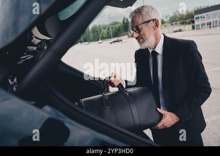 Senior Geschäftsmann im formellen Anzug, der Gepäck in den Kofferraum lädt, sich auf die Reise vorbereitet, draußen auf dem Parkplatz der Stadt Stockfoto