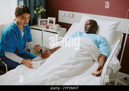 Pflegekraft, die einen älteren Patienten während der medizinischen Untersuchung im Krankenhausbett unterstützt, medizinische Geräte auf dem Betttisch sichtbar, wodurch eine klinische Umgebung geschaffen wird Stockfoto