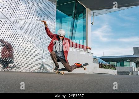 Skateboarder mit Helm und lässiger Kleidung balancieren auf einem Bein, während er einen Trick auf seinem Skateboard vor einem modernen Gebäude ausführt Stockfoto