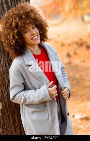 Eine junge Frau mit lockigen Haaren lehnt sich an einen Baum, trägt einen warmen Pullover, lächelt fröhlich im goldenen Herbstlaub und umhergefallenen Blättern Stockfoto