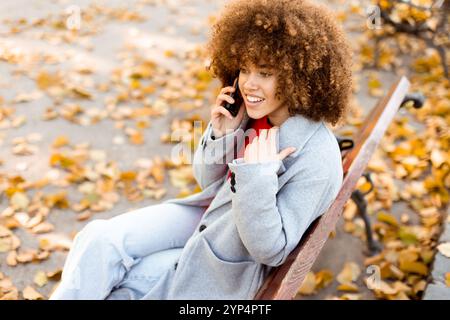 Eine junge Frau mit lockigen Haaren lacht fröhlich, während sie auf einer Parkbank sitzt, die mit lebhaften Herbstblättern bedeckt ist Stockfoto