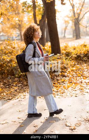 Die fröhliche Frau steht inmitten des bunten Herbstblatts in einem Park, schlürft ein warmes Getränk und trägt ein gemütliches Outfit, das die Schönheit der Saison in der verkörpert Stockfoto