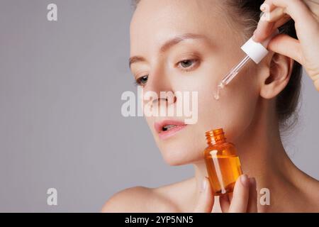 Glückliche lächelnde Frau, die Serum auf ihr Gesicht applyte, die Augen mit Vergnügen schließen. Rotschopf Mädchen hält Dropper mit Hautpflegeprodukt für gesund und glühend Stockfoto