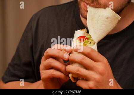 Ein Mann mit einem kleinen Bart beißt in ein Pita-Brötchen, gefüllt mit Fleisch, frischem Gemüse und Salat. Das Foto, das in der Mitte der Bewegung aufgenommen wurde, betont das Deliciou Stockfoto