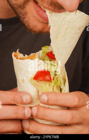 Ein Mann mit einem kleinen Bart beißt in ein Pita-Brötchen, gefüllt mit Fleisch, frischem Gemüse und Salat. Das Foto, das in der Mitte der Bewegung aufgenommen wurde, betont das Deliciou Stockfoto