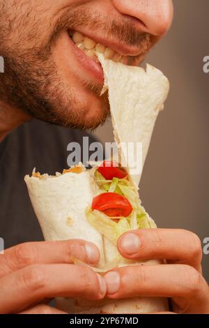 Ein Mann mit einem kleinen Bart beißt in ein Pita-Brötchen, gefüllt mit Fleisch, frischem Gemüse und Salat. Das Foto, das in der Mitte der Bewegung aufgenommen wurde, betont das Deliciou Stockfoto