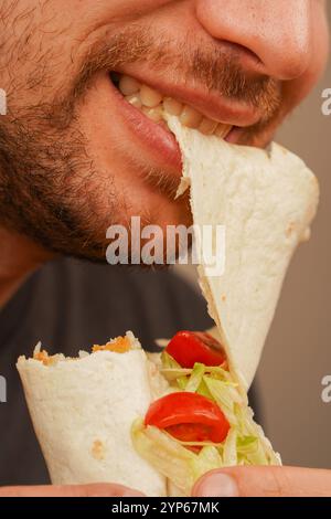 Ein Mann mit einem kleinen Bart beißt in ein Pita-Brötchen, gefüllt mit Fleisch, frischem Gemüse und Salat. Das Foto, das in der Mitte der Bewegung aufgenommen wurde, betont das Deliciou Stockfoto