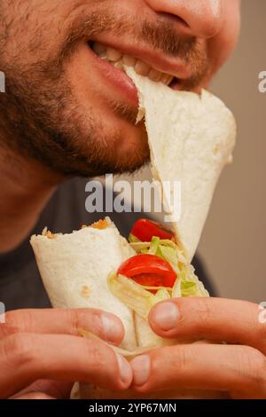 Ein Mann mit einem kleinen Bart beißt in ein Pita-Brötchen, gefüllt mit Fleisch, frischem Gemüse und Salat. Das Foto, das in der Mitte der Bewegung aufgenommen wurde, betont das Deliciou Stockfoto