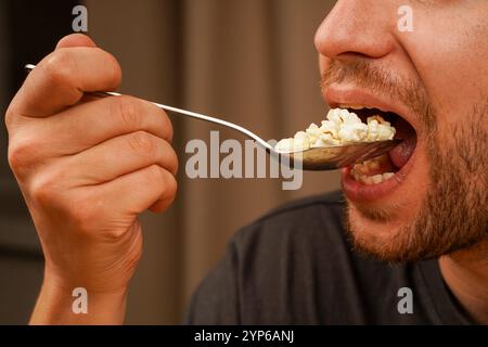 Ein Mann ist dabei, eine Handvoll Popcorn in einem Löffel zu genießen, wobei er sich auf die Details des Imbisses und den Moment der Mahlzeit konzentriert. Ideal für Snacks oder Unterhaltung Stockfoto